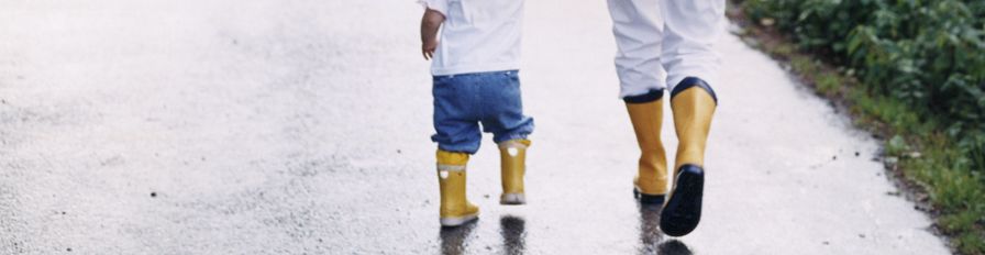 A child and an adult both wearing rain boots walking on the sidewalk after a rainstorm